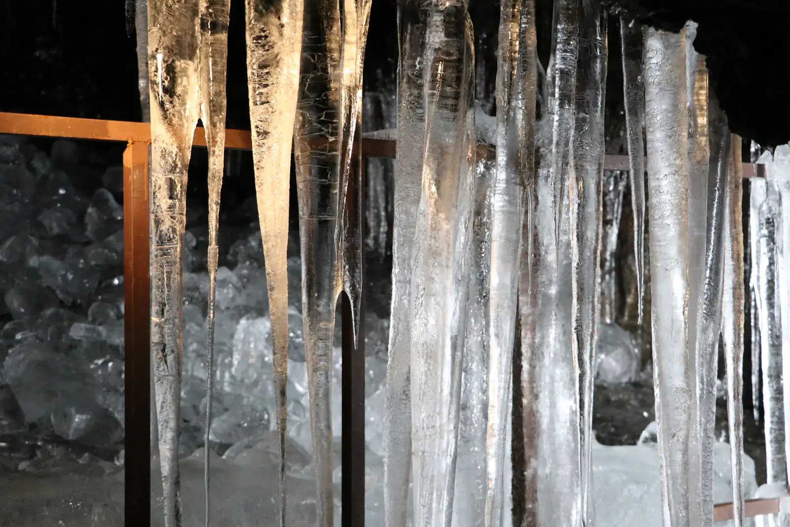 Long clear icicles hanging from the dark rock roof of Narusawa Ice Cave, with blocks of ice stacked behind a railing